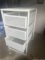 White metal organizer with three mesh drawers, set on a concrete surface with partial garage or workspace background.