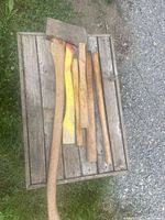 Six vintage axe heads and six wooden axe handles laid out on a wooden surface outdoors.