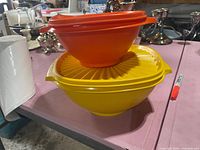 Two vintage Tupperware bowls, one orange and one yellow, stacked with their lids, placed on a table surrounded by various items.