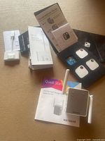 Image showing Netgear AC750 WiFi Extender with quick start guide, three Tile trackers (two Tile Mate and one Tile Slim), wireless CarPlay adapter, packaging and accessories on a beige surface.