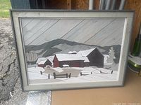 Frontal photo of a wooden folk art landscape showing snow-covered barns and mountains in a gray wooden frame with glass