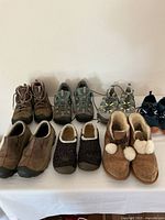 Seven pairs of women's shoes displayed on a table including hiking boots, hiking shoes, slippers, running shoes, and swimming shoes in various earth tones and colors.