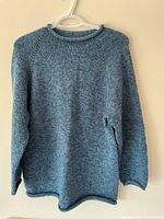 Blue hand knit crew neck pullover sweater hung on white hanger against beige wall, showing knit texture and raglan sleeves.