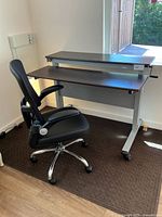Photo showing full adjustable sit-stand desk with dark wood finish surfaces and black office chair on wheels placed in room corner near window.