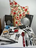 Wide view of all kitchen and baking supplies laid out on a white tablecloth including the printed apron, pans, spatulas, cutting tools and measuring cups.