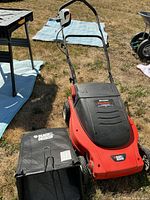 Full view of the red and black electric lawn mower with grass collection bag on dry grass outdoors.
