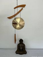 Full view of the hanging gong wind chime with the bronze Buddha sculpture beneath it on a flat surface.