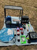 Full lot laid out on blue picnic blanket in grass, showing metal folding table, propane grill box, mosquito tent, sleeping bag, electric lanterns, and small camping accessories