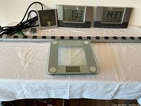 Full view of all items arranged on table: two digital clocks, glass scale, and power strip.