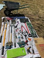 Wide shot showing all gardening tools laid on white cloth on grass with wheelbarrow at rear.