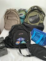 Overall view of seven bags including backpacks, a lunch bag, and a fanny pack arranged on a white surface.