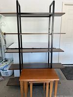 Shelf with black metal frame and five laminate shelves, standing upright in a garage. Nesting tables are placed in front of it.