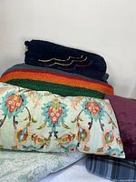 Photo of assortment of soft throw blankets and pillows showing large colorful striped blanket, floral pillows, and other blankets stacked.