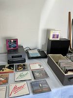 Photo showing Sony compact disc receiver and CD/DVD player along with two black Sony speakers, multiple CDs laid out on table, and fabric bins with CDs and CD holders.