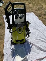 Front view of Sun Joe pressure washer with detergent bottles, spray gun, and power cord visible. Machine placed on white cloth outdoors.