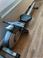 Full view of the rowing machine showing the sliding seat, handlebar, and footrests on a wooden floor near a wall outlet.