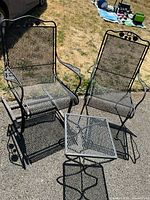 Photo showing two black metal patio chairs, one rocking and one standard, with diamond mesh seating and decorative backrests, alongside small square mesh topped side table.