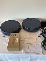 Two black round robot vacuum cleaners on table alongside manuals and a labeled box with Roomba filter