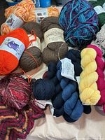 Yarn skeins of orange, brown, multicolor, navy, yellow, and pink wool/acrylic blends, with knitting projects and tools.