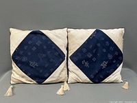 Two Asian throw pillows with cream fabric and central blue embroidered panels featuring Asian characters and tassels at corners.