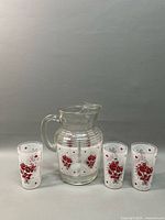 Photo showing clear glass pitcher mug with handle and spout together with three matching tall glass tumblers decorated with red floral and white lattice pattern.