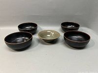 Five Japanese pottery stoneware bowls arranged on a white surface with four black bowls and one beige bowl in the center