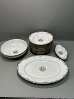 Full set showing 12 stacked soup plates, 2 serving bowls, 1 oval platter, and 1 small dish all with gold trim and central classical medallion artwork.