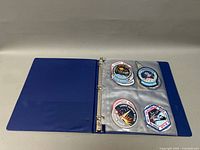 Blue binder open showing four space mission patches in clear plastic sleeves: two on left and two on right.