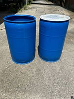 Two blue plastic drums standing on paved ground outdoors, one with lid and clamp, one open-topped.