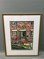 Framed watercolor painting showing front porch with red brick wall, flowers, door, windows, artist signature visible.