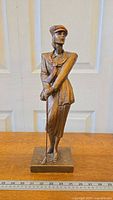 Full front view of the bronze-colored lady golfer sculpture holding a golf club with a square base, placed on a wooden surface in front of white paneled doors.