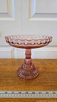 Photo of Atterbury Pink pedestal dish on wooden surface with white doors background, showing bubble detail on rim and fluted pedestal base with measuring scale in front.