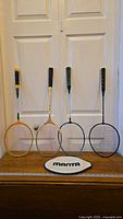 Photo showing four badminton rackets standing upright with two wooden-framed rackets on left and two metal or composite rackets on right, plus Manta oval carrying case on table in front.