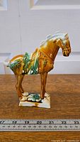 Full view of the horse statue next to a ruler showing length of about 7 inches and height around 6.5 inches. The statue is glazed in brown, green, and cream colors.
