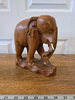 Front left angle view of the handcarved wooden elephant showing detailed trunk and tusks, with a measuring tape for scale.
