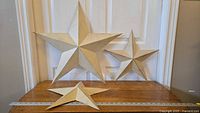 Overall view of three distressed cream-colored metal stars of varying sizes displayed on wooden surface against white door. Two smaller stars approximately 16 inches and one larger star approximately 24 inches.