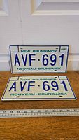 Pair of New Brunswick license plates from 1987 with serial AVF 691 and bilingual text, on a wooden surface with ruler for scale.