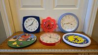 Six vintage clocks placed against a door, showing variety in frame shapes and face designs.