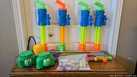 Four water blaster toys, hand pump, two green frog-shaped water containers, assorted sand molds, and bag of chalk laid out on a wooden surface in front of a door.