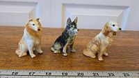 Three glazed ceramic dog figurines side by side on wooden surface with ruler showing size about 3 inches tall. Includes collie, German shepherd, and spaniel-type dog.