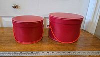 Front view of two round red storage boxes with orange-red rope handles placed side by side on a wooden surface, showing their sizes and lids in excellent condition.