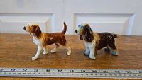 Both ceramic dog figurines shown on wooden surface with measuring tape for size reference, about 4-5 inches tall.