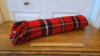 Rolled up red plaid Faribo acrylic throw blanket next to a wooden ruler on a wooden surface.