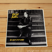 Front cover of the Billy Joel 'An Innocent Man' vinyl LP showing Billy Joel sitting on metal stairs in black and white with yellow font title.