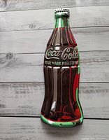 Front side of the Coca-Cola tin bottle sign with embossed and colored details resembling the iconic Coke bottle.