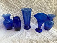 Five assorted blue glass vases shown on white quilted background, varying shapes and sizes