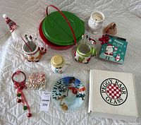 Photo showing all assorted Christmas holiday tableware and decor items including mugs with toothpicks, candle, glass bowl in box, pie carrier, jingle bell door hanger, and holiday picks.