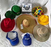 Lot of assorted hats and headwear laid out on white quilted surface including novelty, safari, baseball and party styles with boxed item center back