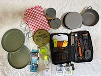 Wide view of all items in the camping mess kit including pans, pot, cups, napkins, cooking utensils, and accessories.
