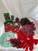 View of all Christmas decorations including bows, placemats, garlands, artificial flowers, and rosette ribbons laid out on a white quilted textile surface.